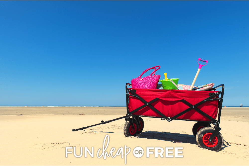 Image of a bright red utility wagon on a sandy beach with a blue sky in the background. The wagon is full of beach toys. 