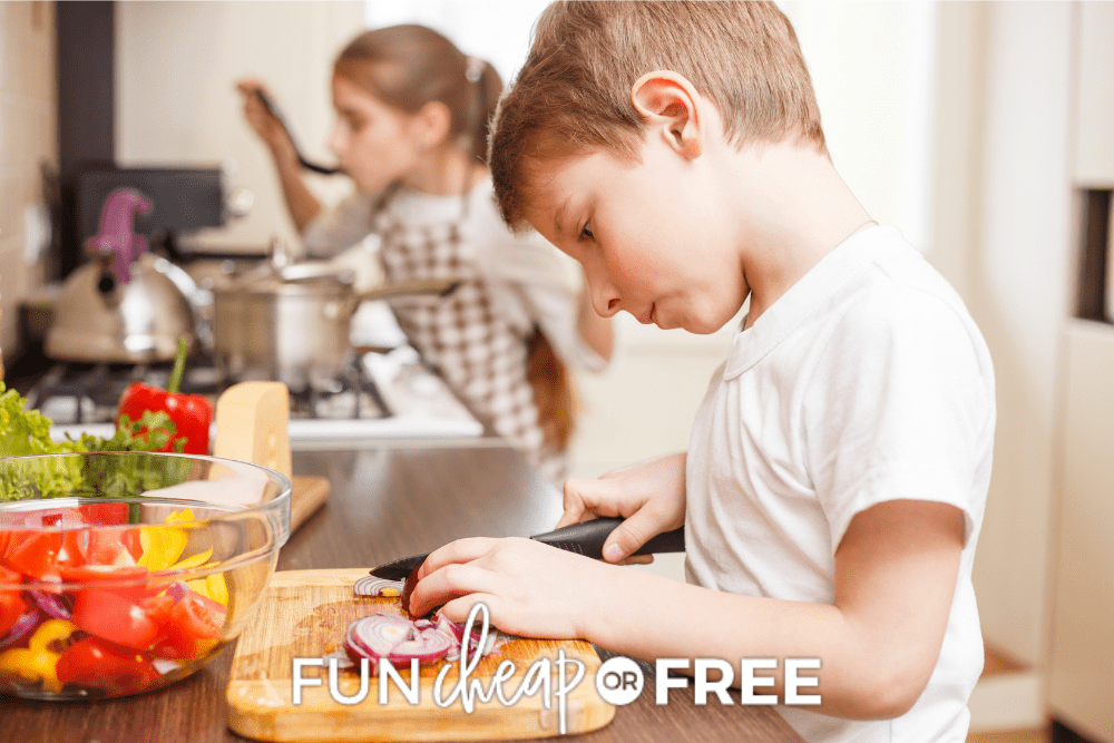 Image of a young boy slicing a red onion on a cutting board for a successful summer routine. In the background a girl is tasting food at the stove. - Fun Cheap or Free