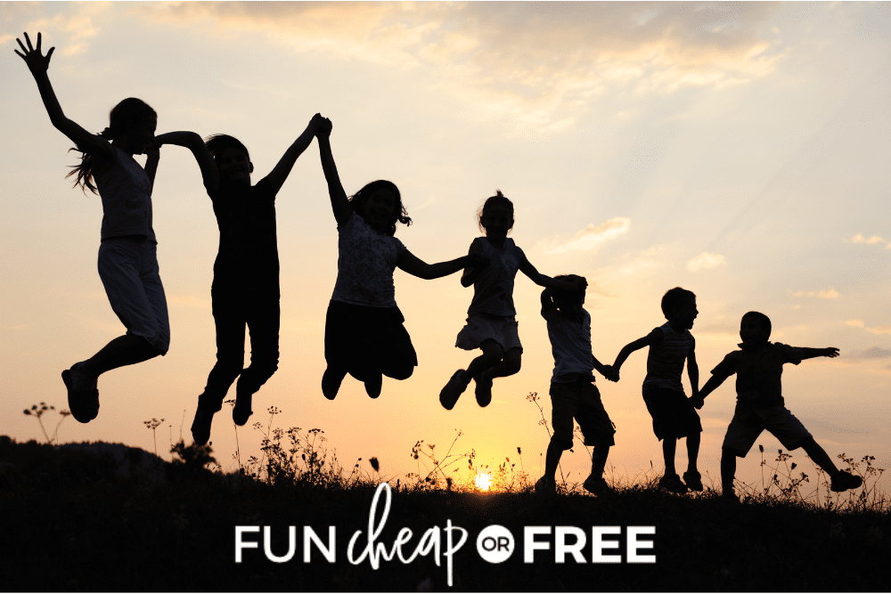 Image of seven children in a meadow silhouetted against the setting sun. They are holding hands and jumping while playing outdoor night games. - Fun Cheap Or Free