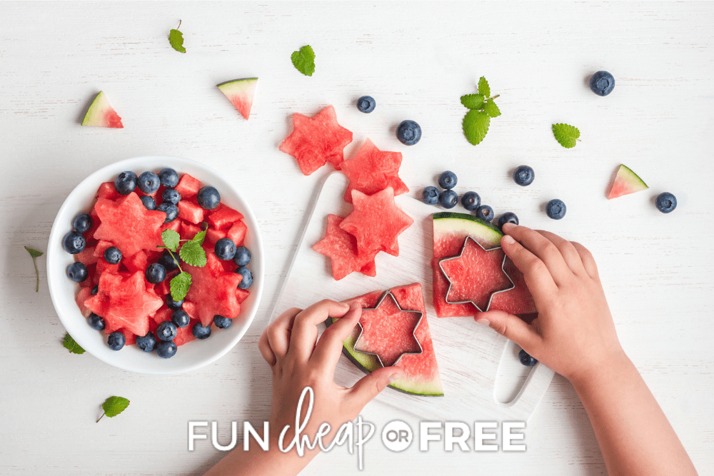 Overhead image of a child using star shaped cookie cutters to cut shapes out of watermelon on a white cutting board. Watermelon shapes and blueberries are in a white bowl next to the cutting board. - Fun Cheap or Free