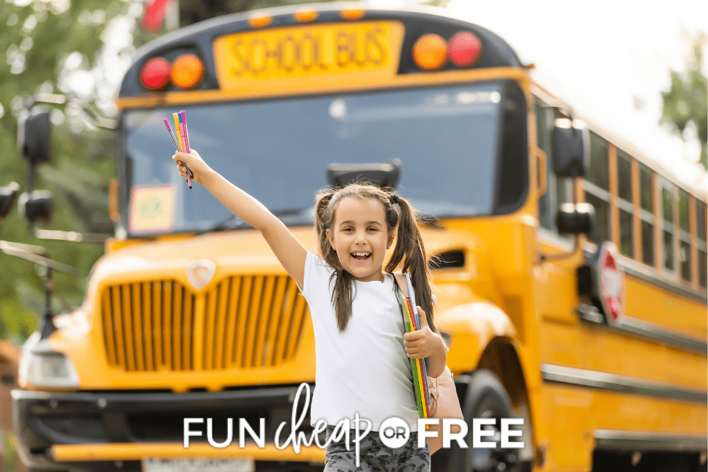 Image of young girl smiling and holding school supplies. She is standing in front of a yellow school bus. - Fun Cheap Or Free