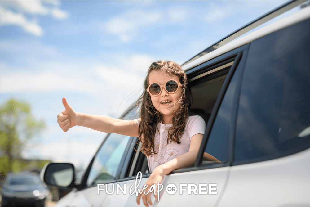A young girl wearing sunglasses is giving a thumbs out out the window of a van during a road trip.