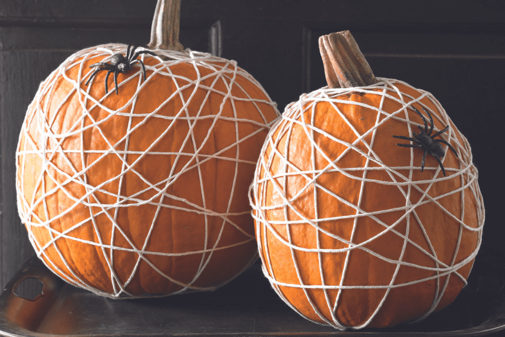 Two orange pumpkins wrapped in white string to represent a spiderweb. There are artificial spiders on the pumpkins. 